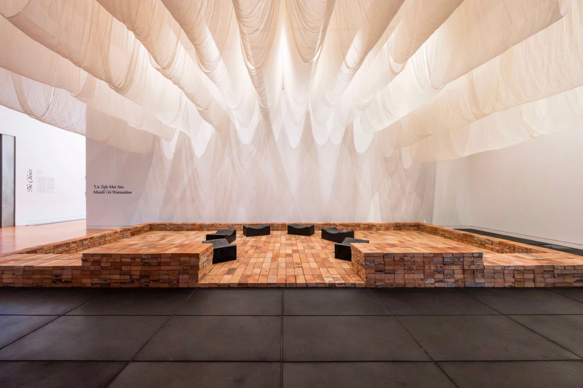 Installation view of a gallery space featuring a large, translucent white fabric canopy suspended from the ceiling, above a raised platform made of reclaimed bricks and timber. Several black sculptural seats are arranged across the platform, with exhibition text on the wall behind.